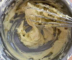 A metal whisk in a bowl of pale yellow, fluffy butter mixture.