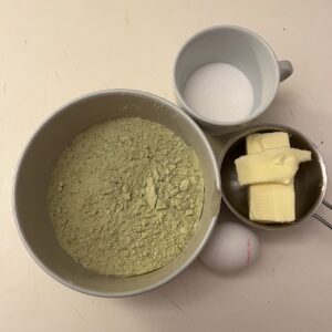 A top-down view of matcha cookie ingredients: a large bowl with matcha powder mixed with flour and baking powder, a cup of granulated sugar, a small metal bowl with butter, and a whole egg.