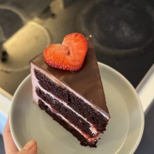 a slice of 3 layer strawberry chocolate cake with pink strawberry frosting and a piece of heart-shape strawberry slice on top of chocolate ganache.